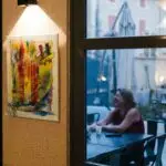 Tableau coloré de Sabine Noël accroché dans un espace intérieur éclairé, visible depuis une terrasse où une personne discute en soirée