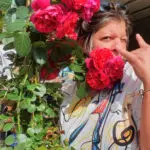 Sabine Noël posant avec humour derrière un bouquet de roses vives, clin d’œil et main levée dans une attitude expressive