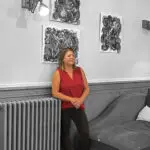Sabine Noël debout dans une galerie d’art, devant ses tableaux en noir et blanc, vêtue d’un haut rouge sur fond désaturé