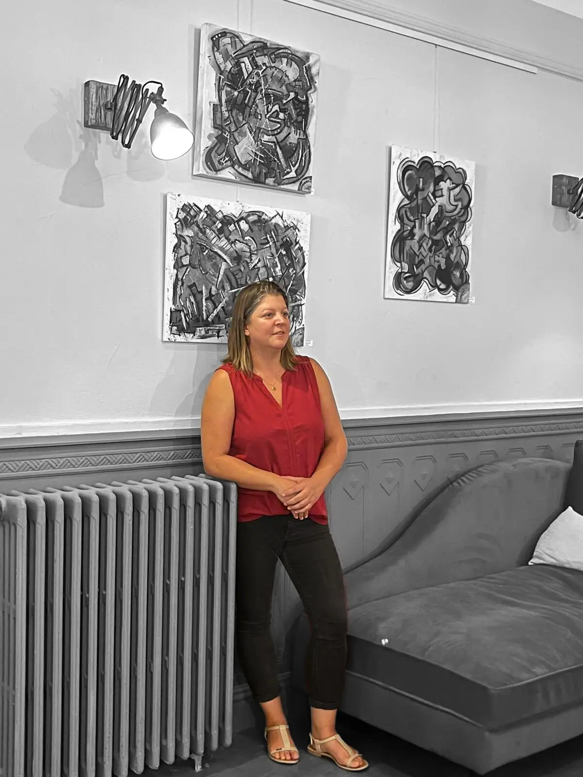 Sabine Noël debout dans une galerie d’art, devant ses tableaux en noir et blanc, vêtue d’un haut rouge sur fond désaturé