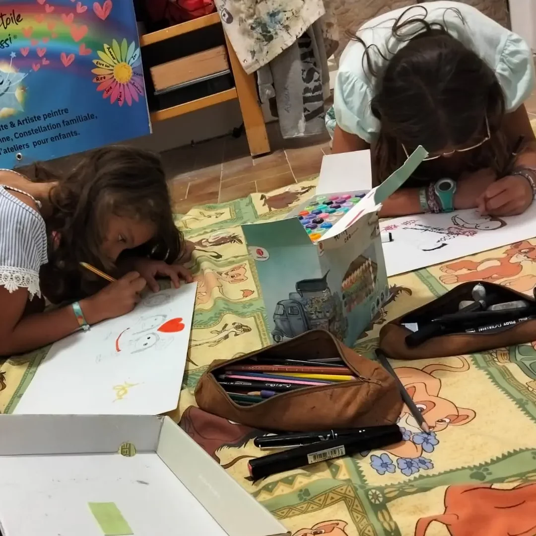 Deux enfants allongés sur un tapis coloré, dessinant et coloriant lors d’un atelier artistique animé par Sabine Noël, art-thérapeute et artiste peintre.